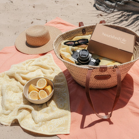 Beach setup with a woven basket, camera, lemons,a hat on a towel and a Nourished Life Beauty Box
