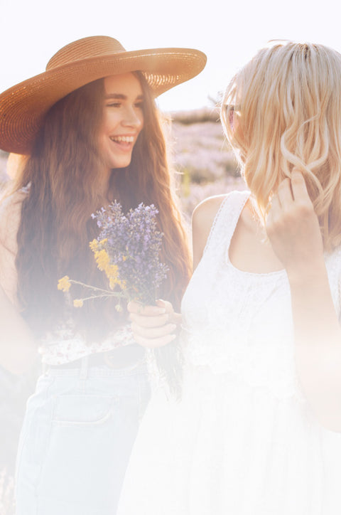 Two women standing close together outdoors, one holding flowers, with a soft, blurred background.