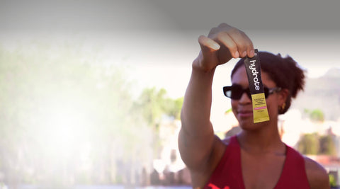 A woman in a red top and sunglasses holds up a small black and yellow Hydroboost energy gel stick pack against a blurred outdoor background.