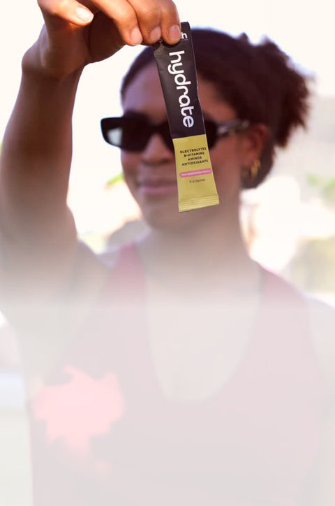 A person outdoors holds up a single Hydrate stick pack in front of their face. The pack is black with white 'hydrate' branding and a yellow lower section.