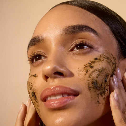 Woman applying a facial scrub to her face with a beige background