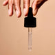 Hand holding black dropper against a tan background.
