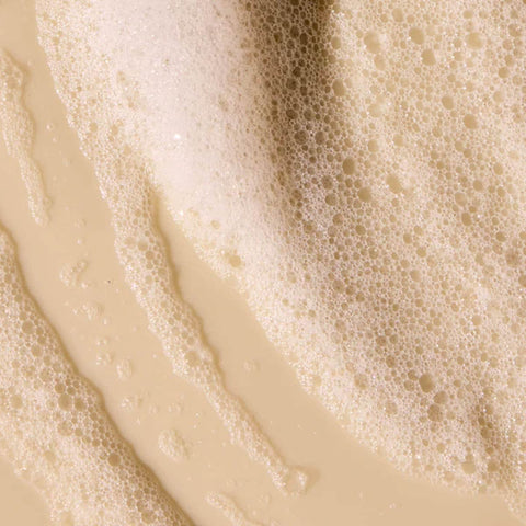 Close up of bubbly shampoo swatch on a beige background.