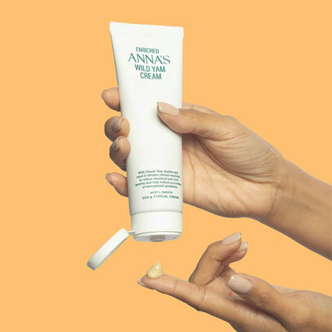 Hand holding a tube of ANNA'S Wild Yam Cream against an orange background.