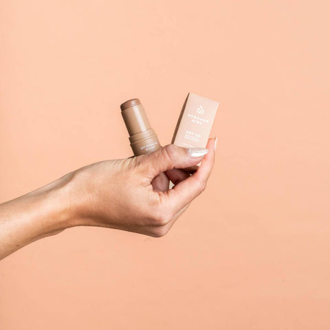 Hand holding a bronzer stick & packaging against a beige background