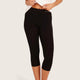 Black capri leggings worn by a model, shown from the waist down against a light beige studio background. The model is wearing a white top and pulling the waistband slightly outward with both hands.
