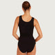 A woman viewed from the back wearing a black sleeveless bodysuit or leotard, standing against a plain light gray studio background.