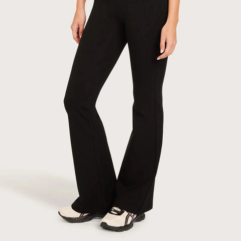 Black flared pants worn by a model, paired with cream and black sneakers, shown from the waist down against a light gray studio background.