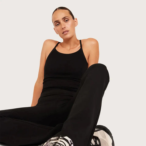 A woman wearing a black tank top and black pants, sitting against a light gray studio background.