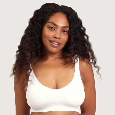 A woman with long, curly dark hair wearing a white wireless bralette, photographed against a solid light gray background.