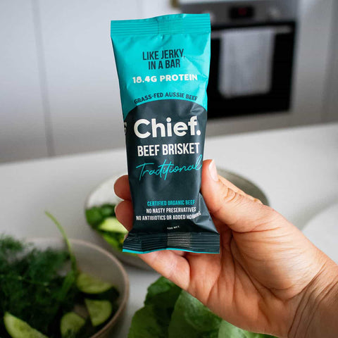 Hand holding a Chief Beef Brisket protein bar package in a kitchen setting.