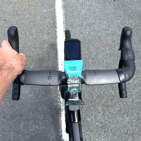 Bicycle handlebar with a package of Chief Beef Jerky attached, on a road background.