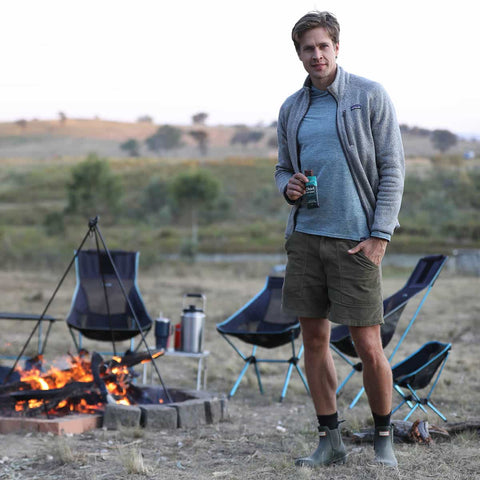 Man standing by a campfire holding a Chief beef bar, with camping chairs and equipment in the background.