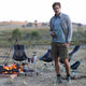 Man standing by a campfire holding a Chief beef bar, with camping chairs and equipment in the background.