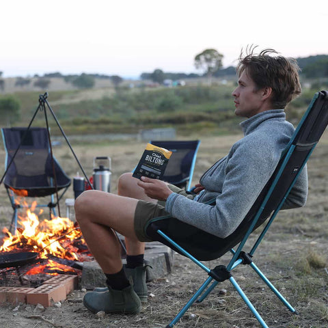 Person sitting by a campfire holding a pack of Chief Biltong Smokey BBQ in a camping chair.