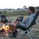 Person sitting by a campfire holding a pack of Chief Biltong Smokey BBQ in a camping chair.