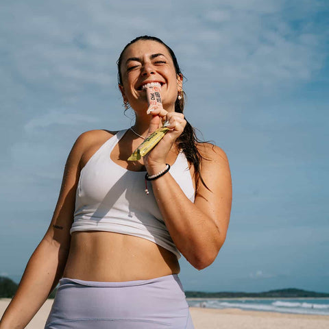 Woman holding a chief collagen bar and biting into the end of it with a happy expression on a beach.