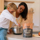 Woman and child in a kitchen with a bag of Chief Beef Liver Powder on the counter while stirring something in a metal bowl.