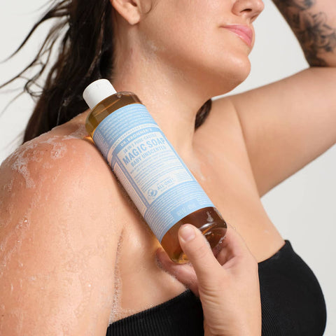Person applying a bottle of 'Dr. Bronner's 18-in-1 Pure-Castile Magic Soap - Baby Unscented' product to their shoulder against a neutral background.