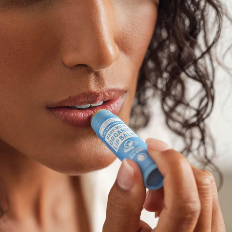 Woman applying a blue Dr Bronner's Baby-Mild Organic Lip Balm to her lips with a blurred background.
