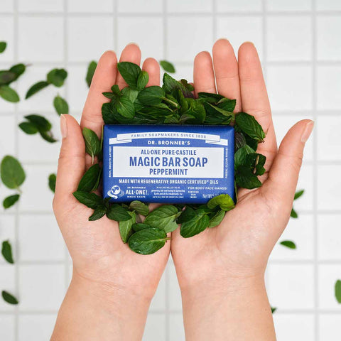 Bar of soap with a blue label held between two hands surrounded by mint leaves on a white tiled background