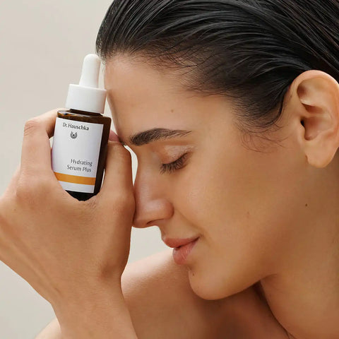 A small amber dropper bottle labeled Dr. Hauschka Hydrating Serum Plus stands beside a lemon and a long root on a clean light gray studio background.