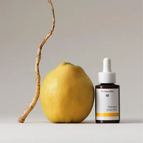 A close-up lifestyle shot of a woman holding a bottle of Dr. Hauschka Hydrating Serum Plus to her forehead, showing the product bottle against her skin.