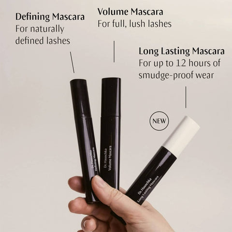 Three Dr. Hauschka mascara tubes held by a hand with product descriptions on a beige background.