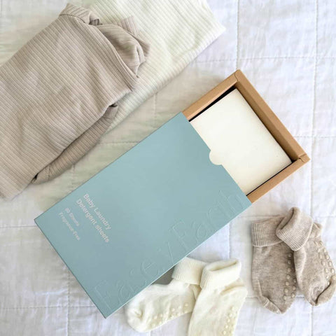 Baby clothes and items including a shirt, socks, and baby laundry detergent sheets on white bedding.