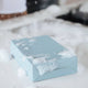 Light blue box of dishwashing detergent sheets with soapy bubbles on a white kitchen counter.