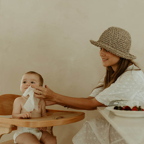 Woman using wipe on baby in a high chair, against a plain background. 