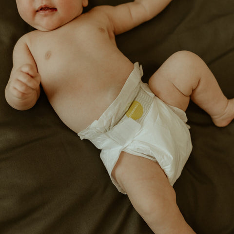 Baby wearing a white nappy on a dark olive bedspread.