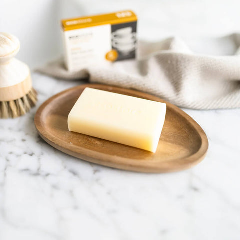 Bar of soap on a wooden plate with a brush and box in the background.