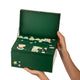 A dark green box held open by two hands against a solid white background. The box features multiple illustrated compartments with numbers and festive designs.