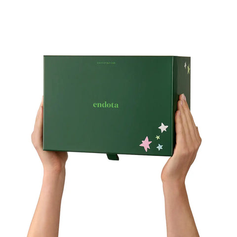 A person holds a dark green Endota product box against a plain white background. The box features the brand name 'endota' in light green, decorated with small pastel-coloured stars on the lower right corner.