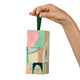 A hand holding a tall, rectangular Endota perfume box with an abstract pastel painting design and a dark green hanging loop, set against a solid white background.