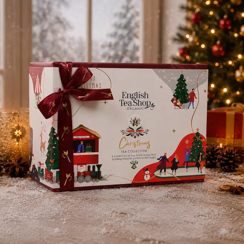 A Christmas tea collection gift box decorated with a dark red ribbon bow and festive illustrations of winter scenes, set on a snowy surface with a blurred Christmas tree in the background.