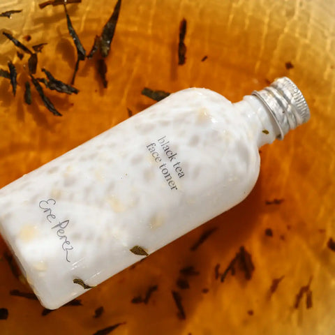 White bottle of Black Tea Face Toner in an orange liquid, surrounded by tea leaves.