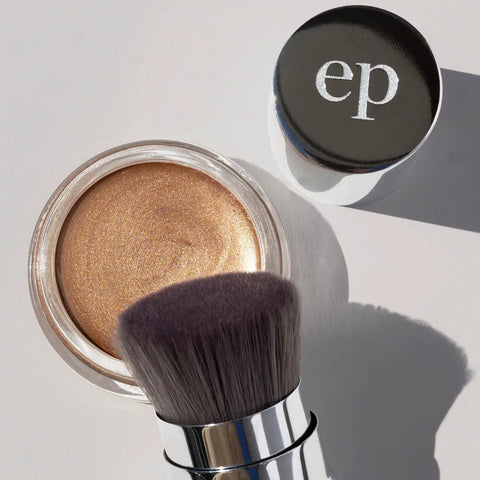 A close-up of a cosmetic product showing a shimmering gold cream makeup in a clear round pot, a makeup brush with dark nylon bristles, housed in a retractable aluminium tube. 