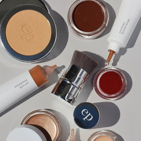 An overhead flat-lay of several cosmetic products on a light gray background, including a pressed powder compact, cream products in squeeze tubes, small open jars of cream products, and a makeup brush. 