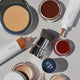 An overhead flat-lay of several cosmetic products on a light gray background, including a pressed powder compact, cream products in squeeze tubes, small open jars of cream products, and a makeup brush. 