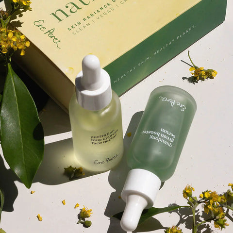 Two serum/oil bottles with droppers next to packaging on a light surface with green leaves and flowers.