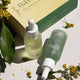 Two serum/oil bottles with droppers next to packaging on a light surface with green leaves and flowers.