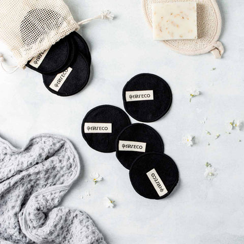 Black makeup removal pads styled on a grey surface spilling out of cotton drawstring bag artistically. Scattered white flowers, soap bar & exfoliator with a towel in the background.