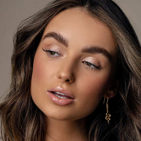 Close-up of a woman with makeup and a gold earring against a neutral background. Wearing Eye of Horus Brow Perfect Dynasty in Medium Brown.