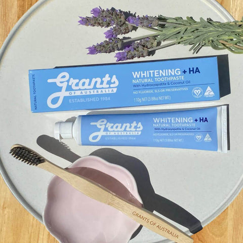 Two tubes of Grants of Australia whitening natural toothpaste on a round grey plate with a wooden toothbrush and lavender flowers.