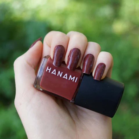 Hand holding a bottle of HANAMI nail polish with matching nail color against a blurred green background