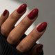 A close-up of a hand with almond-shaped nails painted in a glossy deep burgundy red manicure, wearing a black ribbed sweater.