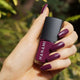 Hand holding a bottle of Hanami nail polish with matching purple nails against a blurred green background