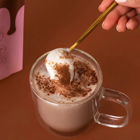 A clear glass mug filled with hot chocolate topped with white foam and cocoa powder, with a hand holding a gold spoon dipping into the foam.